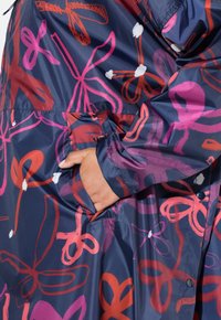 Cappotto impermeabile colorato con sfondo blu navy e audaci motivi floreali rosa, rossi e bianchi, maniche lunghe e tasche laterali.