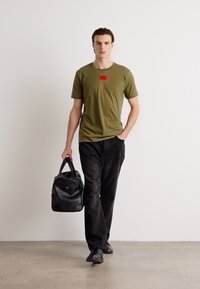 Olíva zöld póló piros, téglalap alakú logóval, fekete nadrággal és fekete bőr táskával párosítva. Slim fit szabás, rövid ujjakkal.