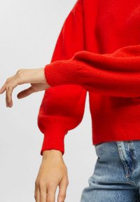 Roter Strickpullover mit geripptem Kragen und Bündchen, Ballonärmeln und einem kurz geschnittenen Design. Kombiniert mit hellblauen Jeans.