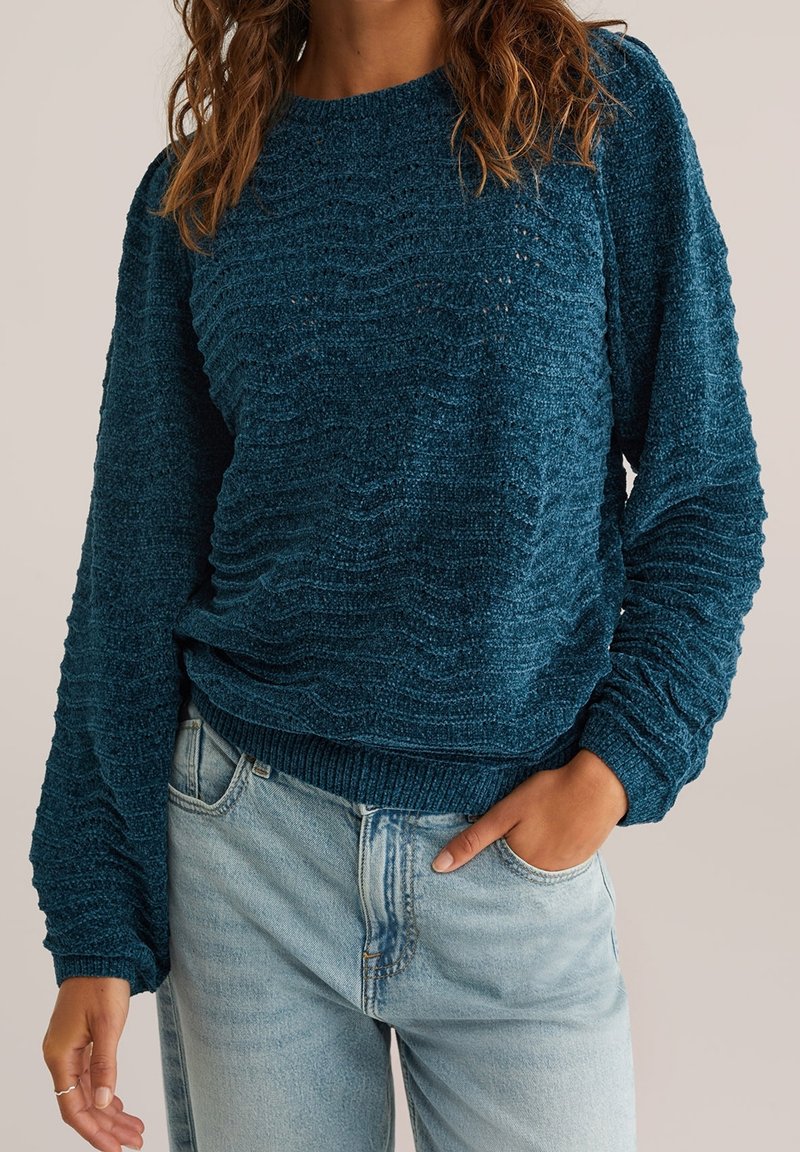 Femme portant un pull texturé bleu sarcelle foncé et un jean bleu clair, une main dans la poche, sur un fond neutre.