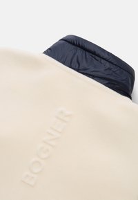 Fleece pullover in cream with a navy nylon collar, featuring embossed brand text on the front, smooth texture, and a relaxed silhouette.