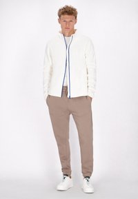 Witte zip-up jas met blauwe details, beige joggerbroeken en witte sneakers. Zachte textuur, casual pasvorm, praktische zakken op de broeken.