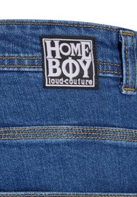 Denim vyötärö, jossa on mustavalkoinen suorakulmainen etiketti, jossa lukee näkyvästi "HOME BOY" ja alla "loud.couture". Keltaiset ompeluyksityiskohdat.