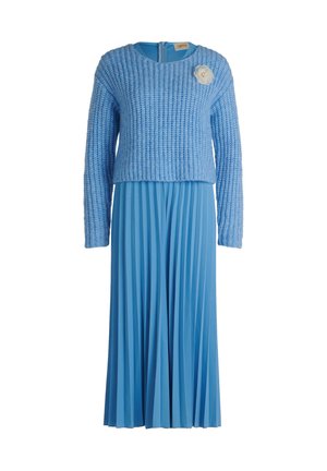 Lyseblå kjole med en tekstureret, strikket kort trøje og plisseret nederdel, fremhævet af en hvid blomstret broche på venstre bryst.