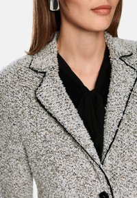 Grau-weiße Blazer mit Struktur und schwarzem Besatz, ausgestattet mit einem Reverskragen und einem Knopfverschluss. Getragen über einem schwarzen Hemd mit einer Krawatte.