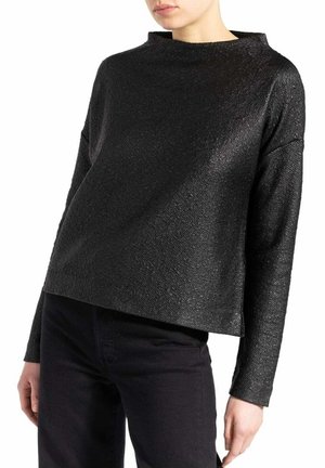 Haut noir à manches longues en tissu texturé avec un col haut et une coupe ample et carrée, associé à un jean foncé.