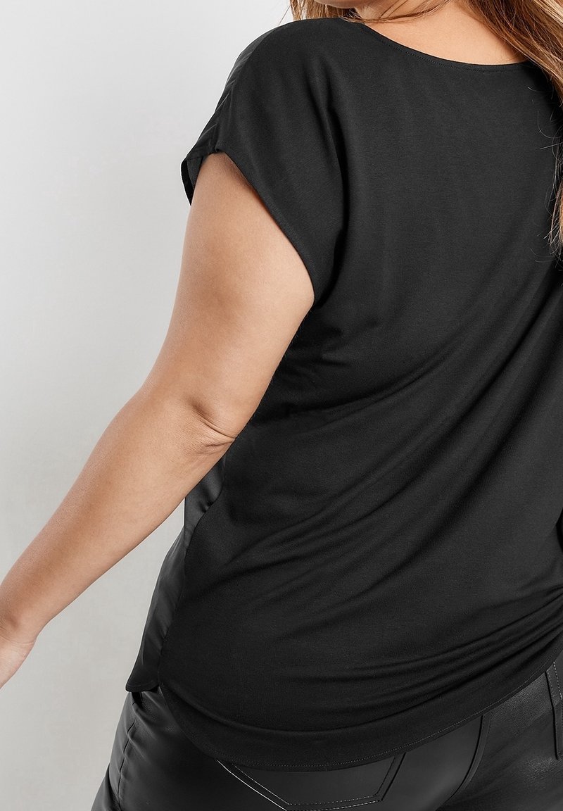 Femme portant un haut noir ample à manches courtes et un pantalon noir, vue de dos et de côté avec le bras tendu.