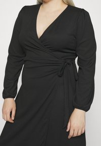 Vestido negro ajustado en tejido texturizado con mangas largas de manga abullonada, escote en V y detalle en la cintura con cinta. Presenta un dobladillo fluido.