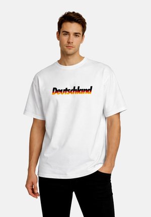 Young man with brown hair wearing a white T-shirt that says "Deutschland" in black, red, and yellow font, standing against a plain background.