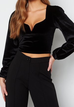Frau trägt ein schwarzes Samt-Cropped-Longsleeve mit herzförmigem Ausschnitt und hoch taillierte schwarze Hose, Hand an der Hüfte.