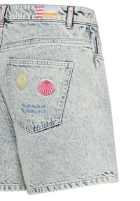 Short en jean bleu clair avec broderie sur la poche arrière représentant un soleil jaune, un coquillage rose et des vagues bleues, accompagné d'une étiquette colorée sur la ceinture.