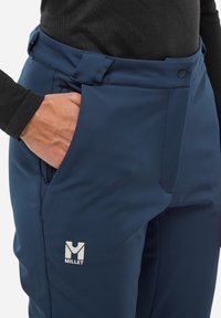 Ręka schowana w kieszeni granatowych spodni outdoorowych marki Millet, noszonych z czarną bluzą z długim rękawem.