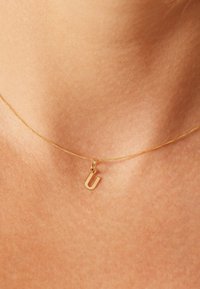 Gouden ketting met een kleine, gepolijste hanger in de vorm van de letter "U". De ketting is fijn en glad, die subtiel licht weerspiegelt.