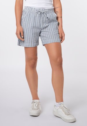 Frau trägt weiße Sneakers und blau-weiß gestreifte Shorts mit Kordelzug, sichtbare Tätowierungen am Unterarm und Oberschenkel.