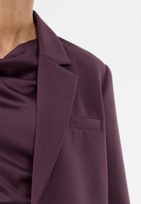 Άτομο που φοράει ένα σκούρο μοβ σακάκι πάνω από μια ταιριαστή μεταξωτή μπλούζα, εμφανίζεται από το λαιμό και πάνω σε έναν απλό φόντο.