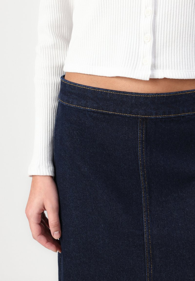 Gestreifte weiße Strickjacke mit Knöpfen, kombiniert mit einem dunklen Jeansrock mit gelben Nähten und geradem Schnitt. Hand liegt auf dem Rock.