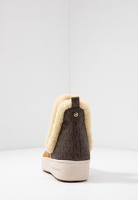 Chaussure marron à motif avec une doublure en beige peluche, arrière court avec un accent de logo doré et une semelle plateforme beige. Matériaux texturés partout.