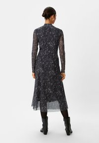 Abito nero a maniche lunghe con una texture trasparente e a motivi; presenta un colletto alto e una lunghezza midi, abbinato a stivali neri fino al ginocchio.