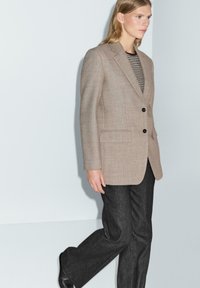 Blazer marrone chiaro, con texture, dotato di due bottoni neri, abbinato a una maglietta a strisce e pantaloni grigio scuro, caratterizzati da un taglio dritto e tasche.