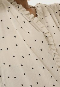 Blouse crème à pois noirs, dotée d'un col à volants et d'un tissu texturé. La coupe est ample avec une silhouette décontractée.