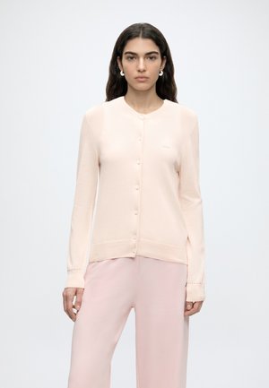 Donna con lunghi capelli scuri indossa un cardigan rosa chiaro abbottonato e pantaloni a gamba larga abbinati, in piedi su uno sfondo bianco uniforme.