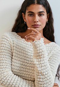 Creme striktrøje med et tekstureret, mønstret design og bølgede udskæringer; modellen har langt hår og rører ved sit ansigt. Sølv ring og hoops øreringe.