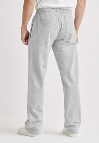 Man draagt lichtgrijze joggingbroek met achterzakken en witte sneakers, staand op een witte vloer.