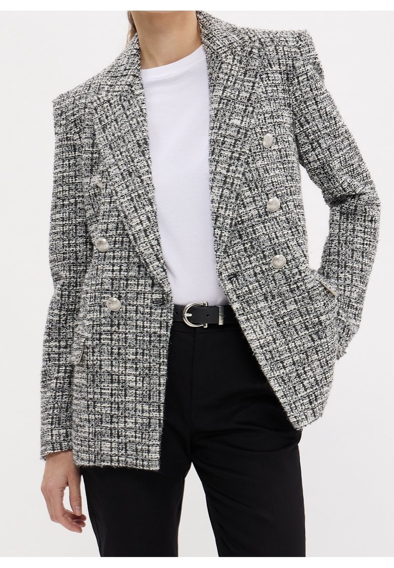 Blazer à carreaux noir et blanc texturé avec design à double boutonnage, boutons argentés et col cranté, porté par-dessus une chemise blanche.
