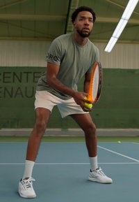 Teniso žaidėjas šviesiai žaliame sportiniame marškinėlyje ir baltuose šortose, laiko teniso raketę ir kamuoliuką mėlyname korte su žaliu fono.
