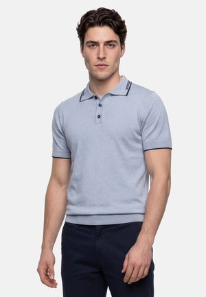 Ung mand med mørkt hår iført en lyseblå kortærmet poloshirt og mørkeblå bukser, stående mod en ensfarvet baggrund.
