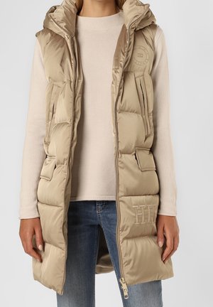 Nainen, joka pukeutunut beigenväriseen toppattuun hihattomaan hupputakkiin vaalean neuleen ja sinisten farkkujen päällä, takissa on vetoketjullinen etuosa ja taskut, joissa on logokoristeet.