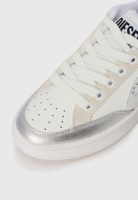 Sneaker bianco con accenti argentati, materiali in suede e pelle, punta perforata, lacci piatti e forma della punta rotonda per un design sportivo.