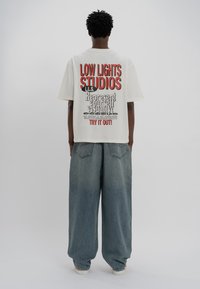 White cotton t-shirt with red and black graphic text on the back, paired with loose-fitting blue denim pants featuring two rear pockets.