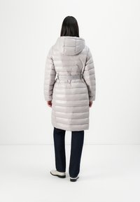 Lysegrå puffjakke med hette, belte i midjen og quiltet design. Modellen har på seg mørkeblå jeans og lyse sko, vist fra ryggen.