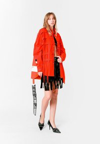 Chaqueta roja de gran tamaño con múltiples bolsillos, top negro corto y falda negra con flecos. Zapatos de tacón alto negros con detalles decorativos.