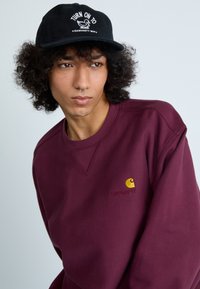 Bordo mikina s kulatým výstřihem a žlutým logem Carhartt na hrudi. Černá kšiltovka s bílým grafickým textem a designem kachny.