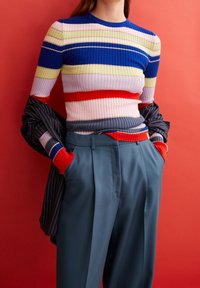 Person klädd i en flerfärgad randig ribbad tröja och blå högmidjade byxor med händerna i fickorna mot en röd bakgrund.