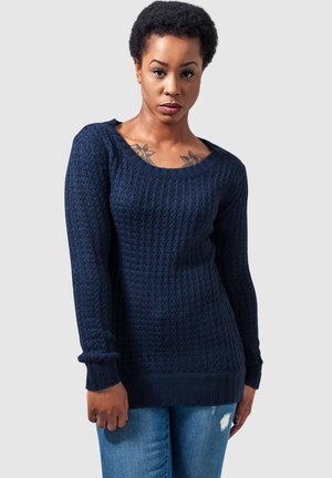Pull en maille bleu marine avec un motif texturé, décolleté large en ras du cou et poignets et ourlet côtelés. Porté avec un jean en denim bleu clair.