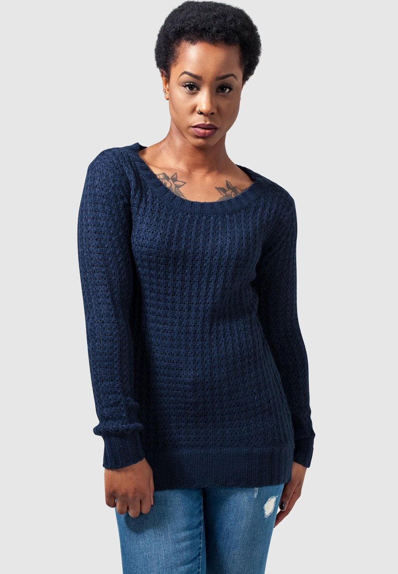 Maglione lavorato a maglia blu navy con motivo testurizzato, ampia scollatura a barchetta e polsini e orlo a coste. Indossato con jeans in denim azzurro chiaro.