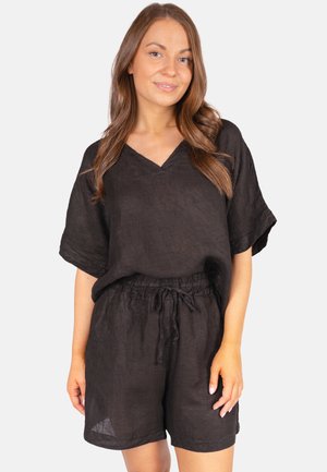Woman with long brown hair wearing a loose black short-sleeve top and matching black shorts against a plain light background.