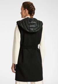 Frau, die steht und einen schwarzen ärmellosen Mantel mit einem taillierten Gürtel und einer gepolsterten schwarzen Kapuze über einem langärmeligen Cremetop trägt, mit dem Rücken zur Kamera.