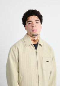 Jachetă crem cu fermoar, cu dungi verticale, având un buzunar pe piept și detalii cu logo. Țesătură moale, cu guler structurat și croială confortabilă.