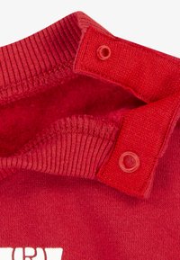 Rød hettegenser detalj som viser en ribbestrikket hals med to trykknapper. Glatt tekstur med en bomullsblanding og trykt logo på forsiden.