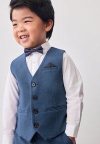 Chaleco azul marino con un acabado texturizado, con botones y un pañuelo de bolsillo, usado sobre una camisa blanca y una corbata de lazo oscura.