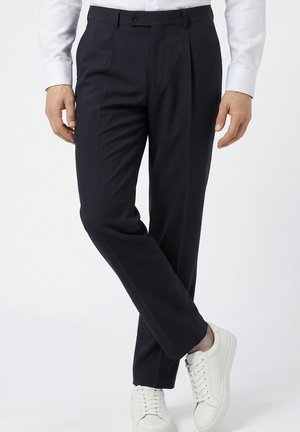 Mann trägt maßgeschneiderte schwarze Hose, weißes Hemd und weiße Sneakers, steht mit gekreuzten Beinen vor schlichten Hintergrund.