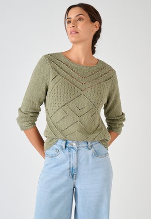 Vrouw met donker haar in een vlecht, gekleed in een lichtgroene gebreide trui met patroon en lichtblauwe spijkerbroek, staand met haar handen achter haar rug.