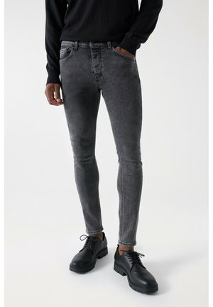 Mężczyzna w wąskich, spranych czarnych dżinsach, czarnym swetrze i czarnych sznurowanych butach, stojący z jedną ręką w kieszeni na białym tle.