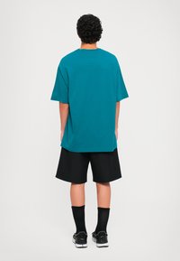 Göndör hajú férfi, akinek teál színű, oversize pólója, fekete nadrágja, fekete zoknijai és fekete sportcipője van, háttal áll egy fehér háttér előtt.