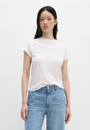 Młoda kobieta z długimi czarnymi włosami, ubrana w zwykły biały t-shirt z krótkim rękawem i jasnoniebieskie, spodnie dżinsowe z wysokim stanem, stojąca na jednolitym tle.
