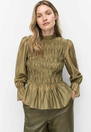 Vrouw met lang blond haar die een olijfgroene geplooide blouse en donkergroene broek draagt, staand met haar handen in haar zakken tegen een witte achtergrond.